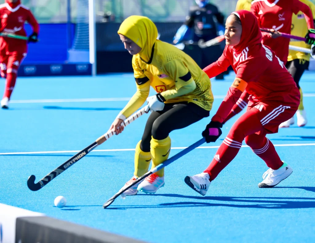 Hockey players in yellow and red kit competing for the ball on a blue artificial pitch