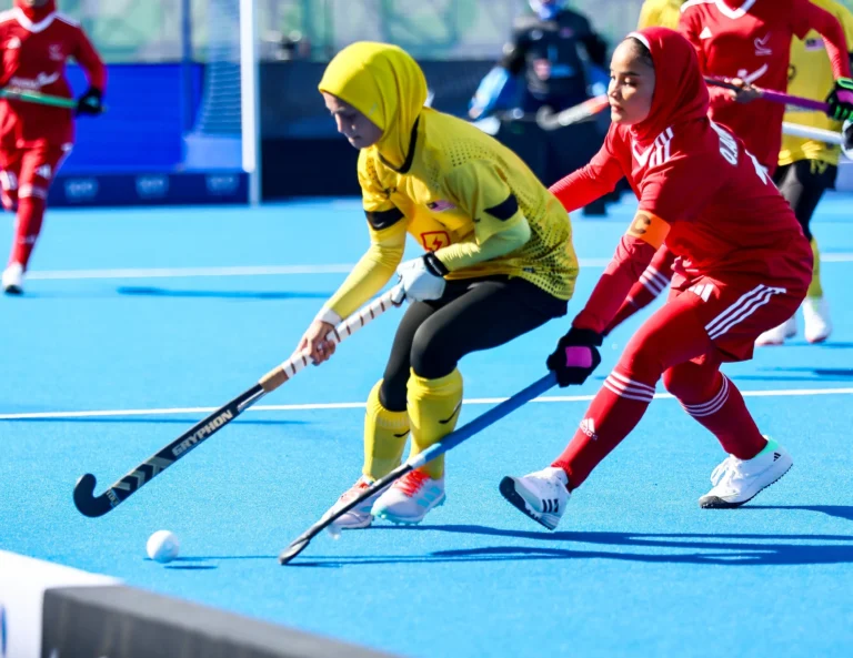Hockey players in yellow and red kit competing for the ball on a blue artificial pitch