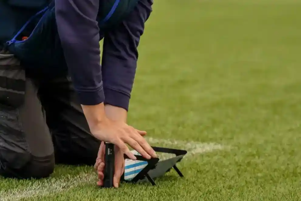 Technician kneeling on a pitch to place a handheld test gauge beside a white line marking