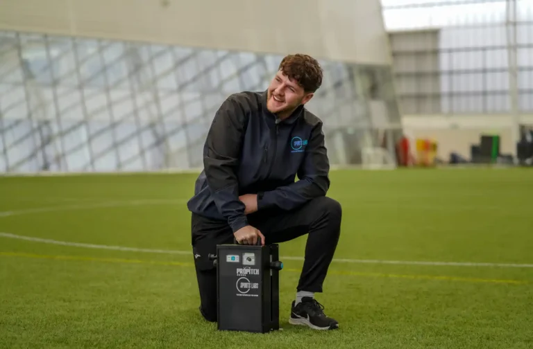 Sports Labs technician seated on a test equipment case on an indoor 3G pitch