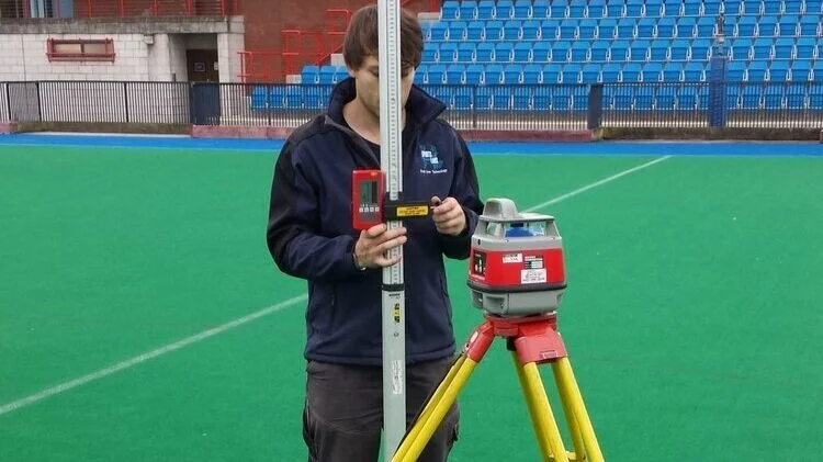 Sports Labs technician using a survey instrument on a tripod beside a stadium pitch