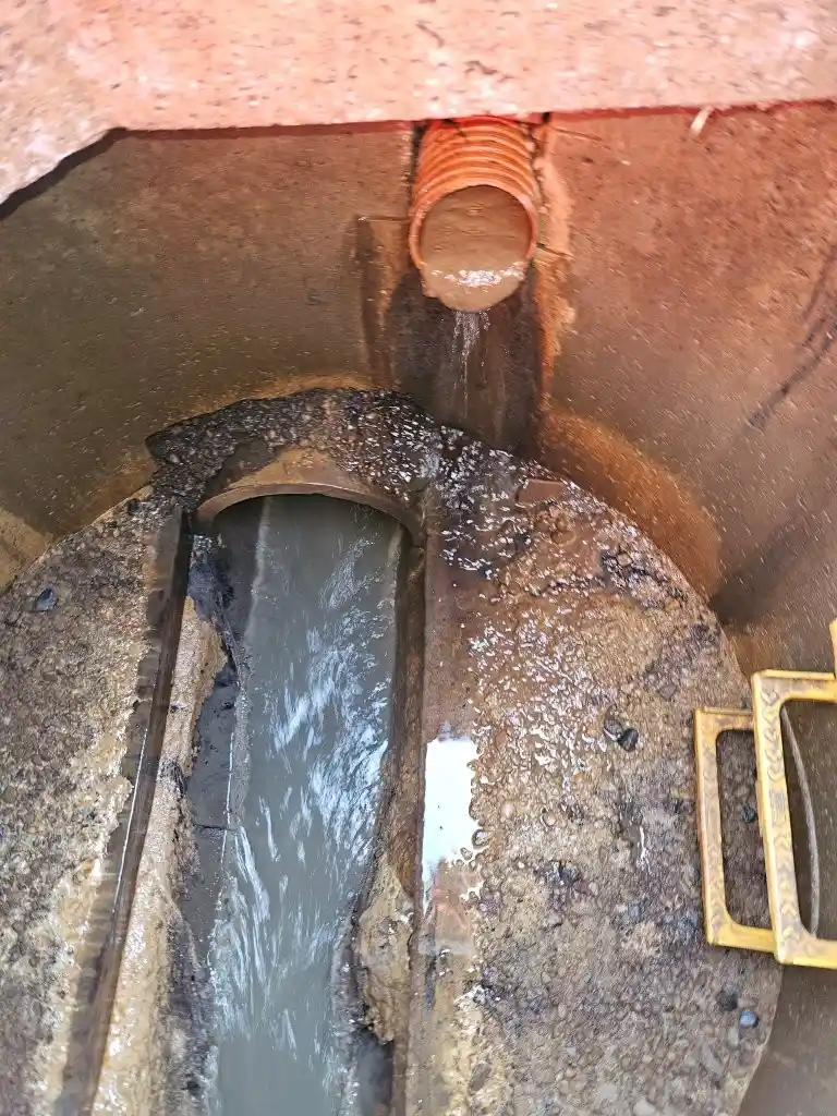 Open drainage manhole beside a sports pitch with a lateral drainage pipe discharging water into the channel