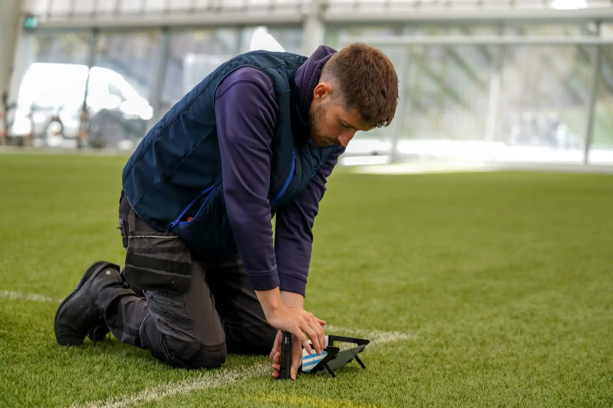 Groundsman kneeling on a natural turf pitch measuring grass length with the Prism Gauge