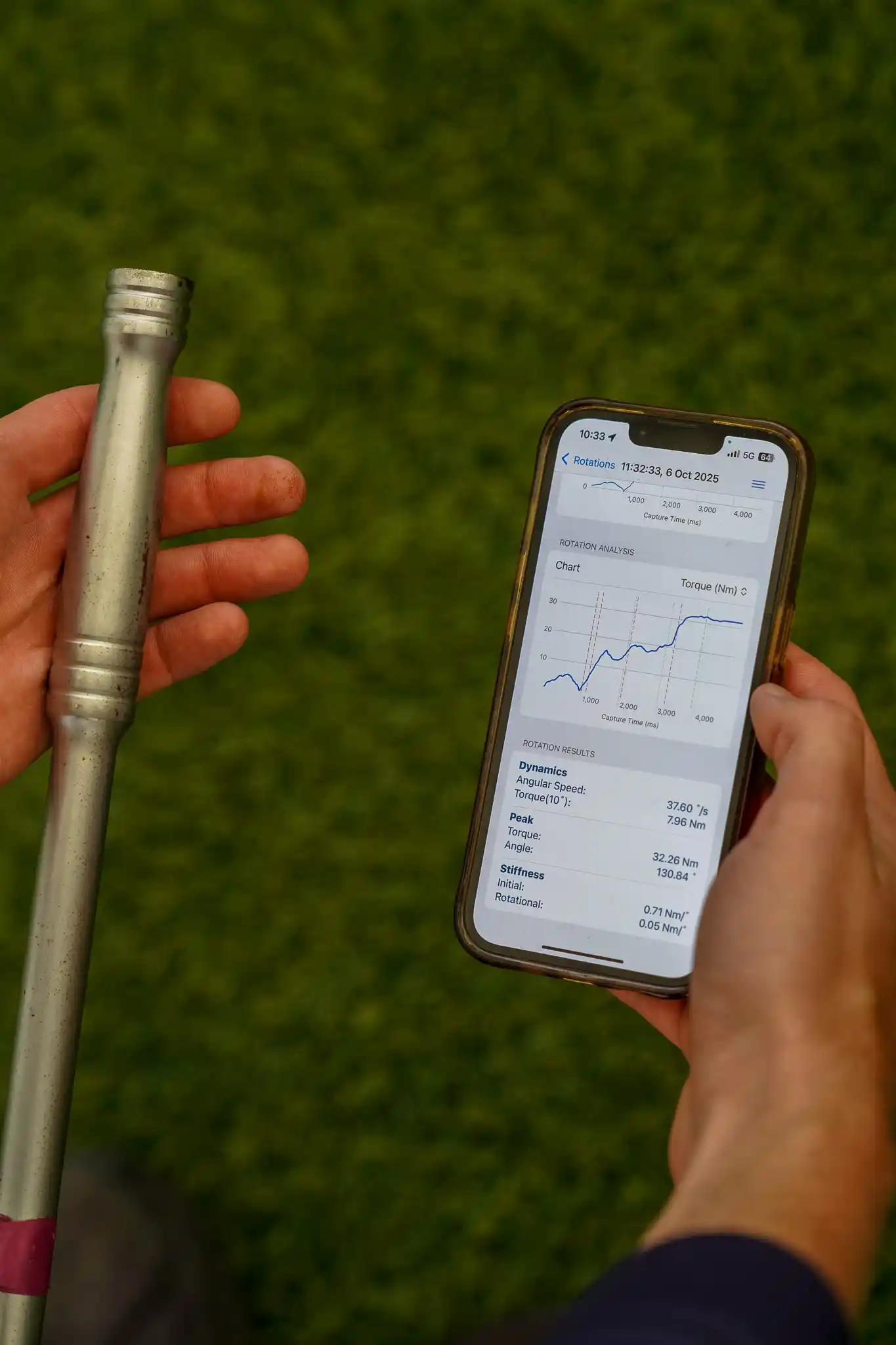 Hand holding a phone showing the ProPitch app beside a test probe on a turf pitch