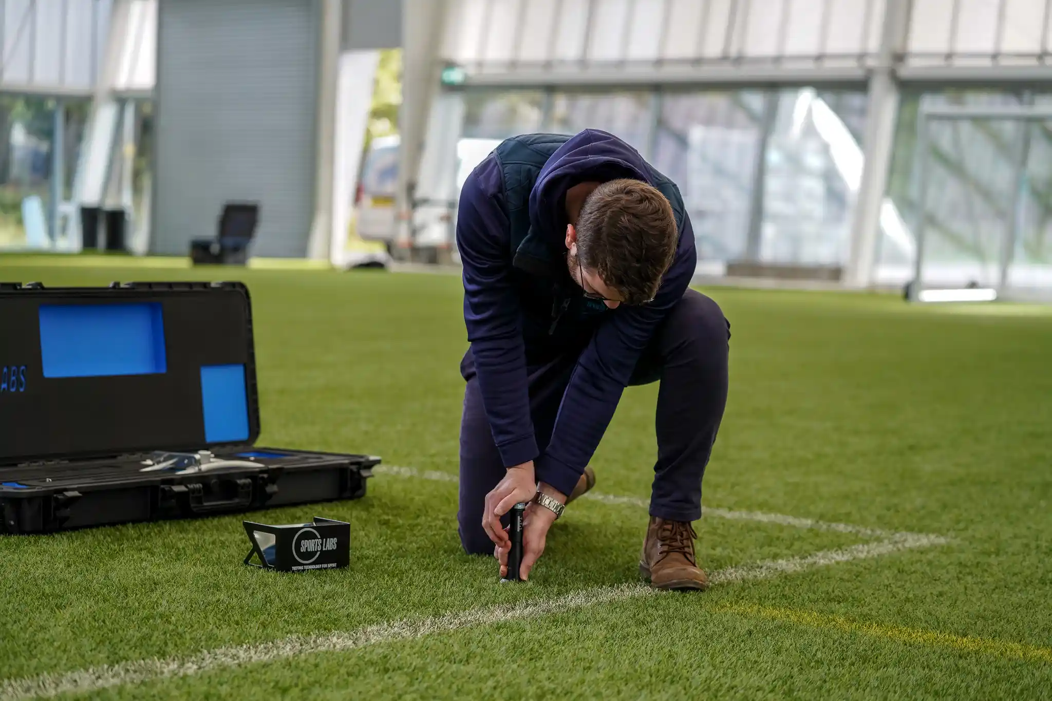 Technician kneeling on an indoor pitch beside an open test case, working along a marked line
